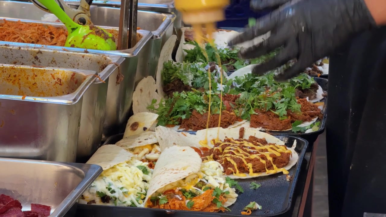 Filmagens em close-up de um vendedor de comida preparando vários tacos e wraps frescos numa chapa quente. Tortillas são recheadas com carne, queijo, vegetais, ervas e molhos durante a movimentada preparação de comida de rua.