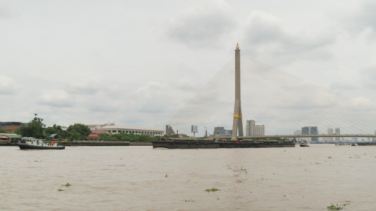 Boats travel along the Chao Phraya River in Bangkok, Thailand, passing beneath a large suspension bridge as traffic and city buildings line the busy waterfront