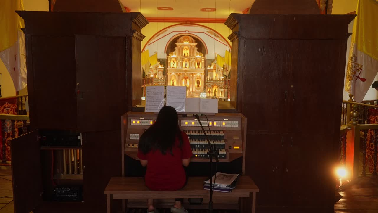 A wider shot of girl playing grand pipe organ in the Minor Basilica, Majayjay, Laguna, Philippines, ornate golden altar glowing behind with rich church details and warm light