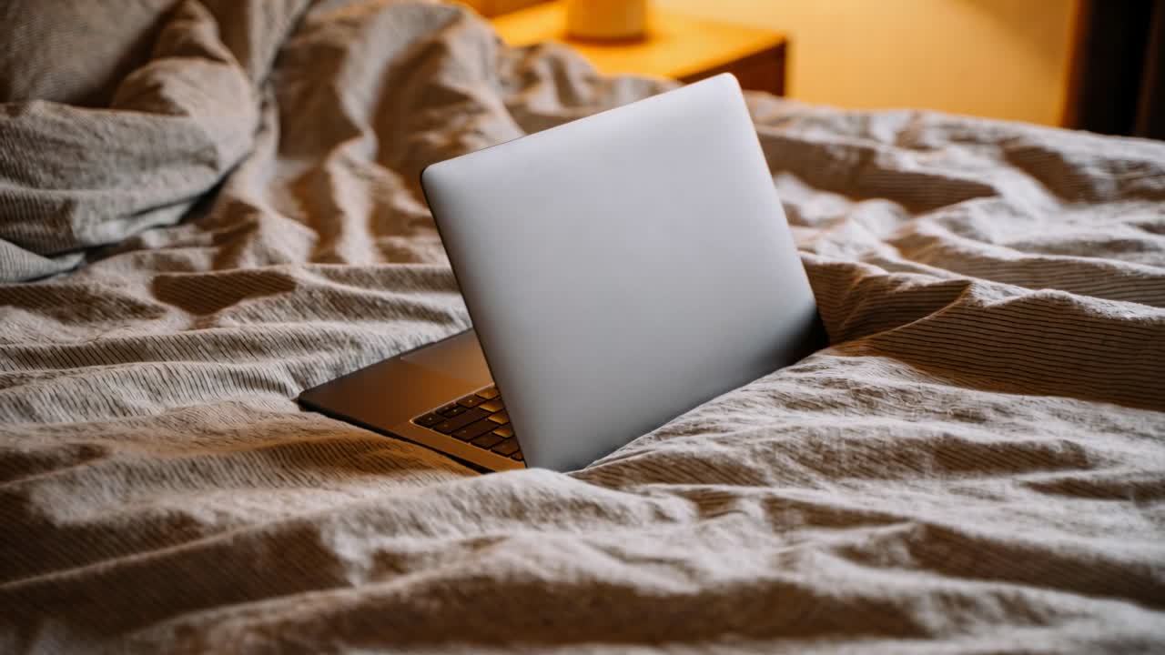 Kamera strammes ind, sølvfarvet laptop ligger halvt åben i krøllet dyne på seng, fremhæver tastatur.