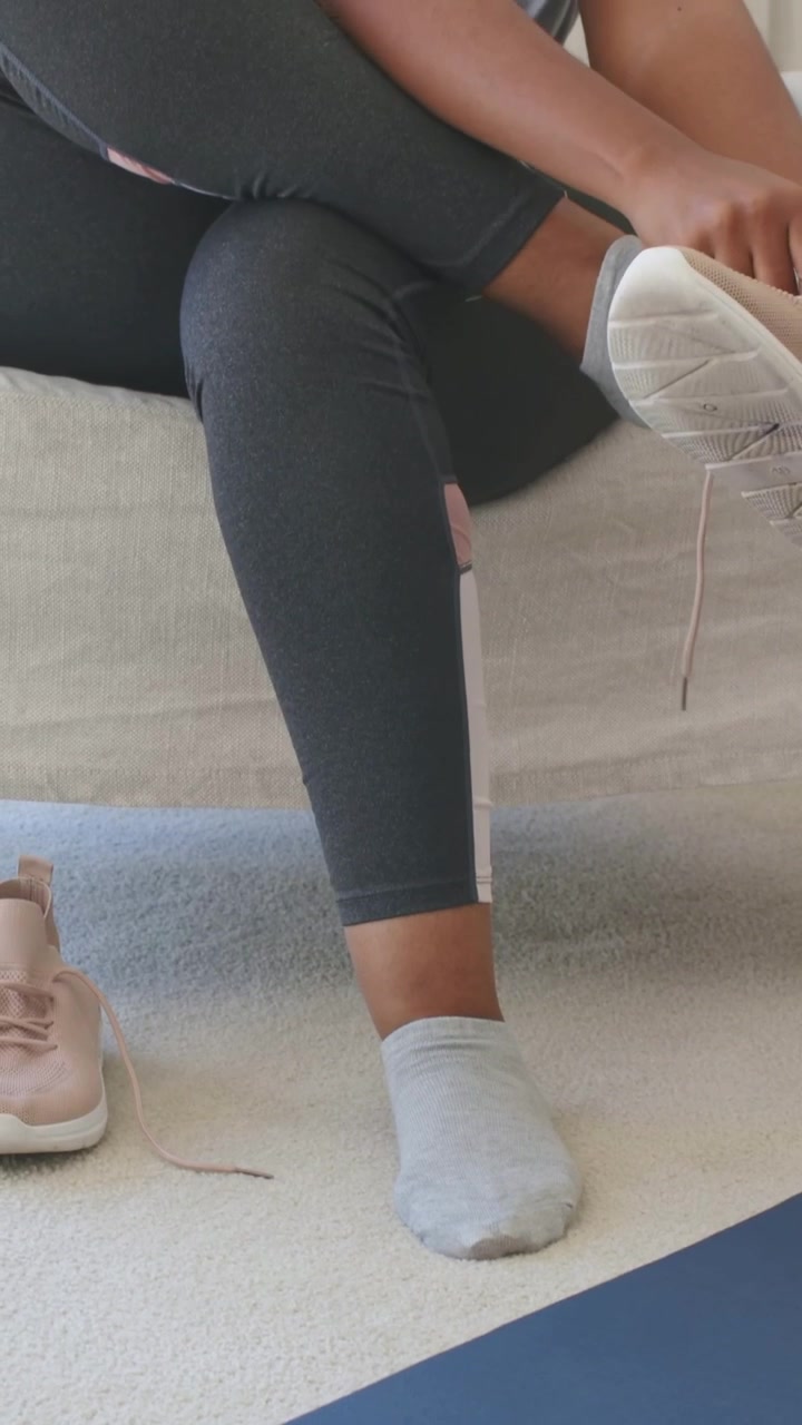 Vertical video: Reaching woman in leggings putting on pink sneaker at home by blue mat for workout