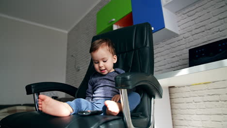beautiful toddler swinging in the big leather chair. child plays with tv remote and saying something. low angle view.