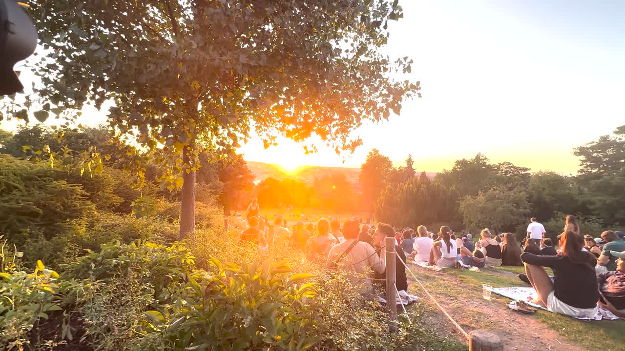 Groups relax on grassy hillside at sunset, camera on tripod, warm summer evening, natural light