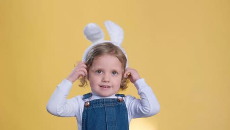 a happy toddler in bunny ears smiles and makes a thumbsup gesture on a yellow background. she plays with her ears, enjoying leisure time and having fun like a little baby