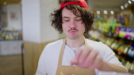 close-up of a confident guy with curly hair and a red cap a supermarket worker chews a bun while doing inventory on the shelves in the supermarket