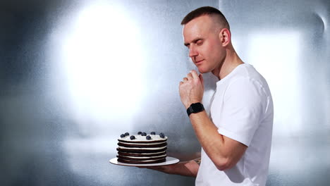 man holding a chocolate cake with blueberries. thoughtful male in white t-shirt looks at dessert and shows it to camera smiling.