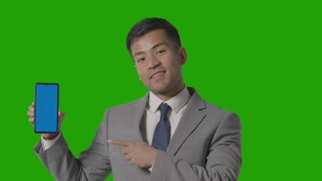 Studio-Shot-Of-Smiling-Businessman-In-Suit-Holding-Blue-Screen-Mobile-Phone-Against-Green-Screen-