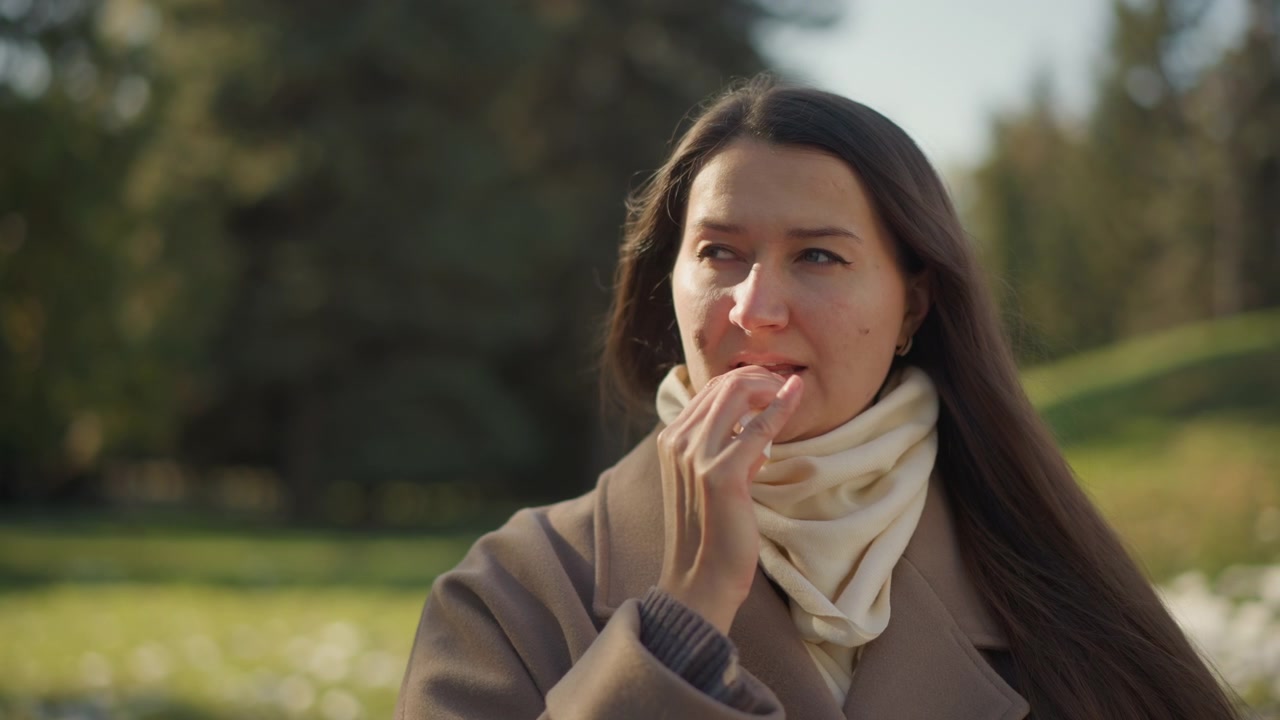 Primo piano di una donna all'aperto, in luce naturale, che indossa un cappotto marrone e una sciarpa color crema. La sua mano è vicino al viso con movimenti sottili. Lo sfondo presenta un paesaggio di parco verde con alberi e una luce soffusa.