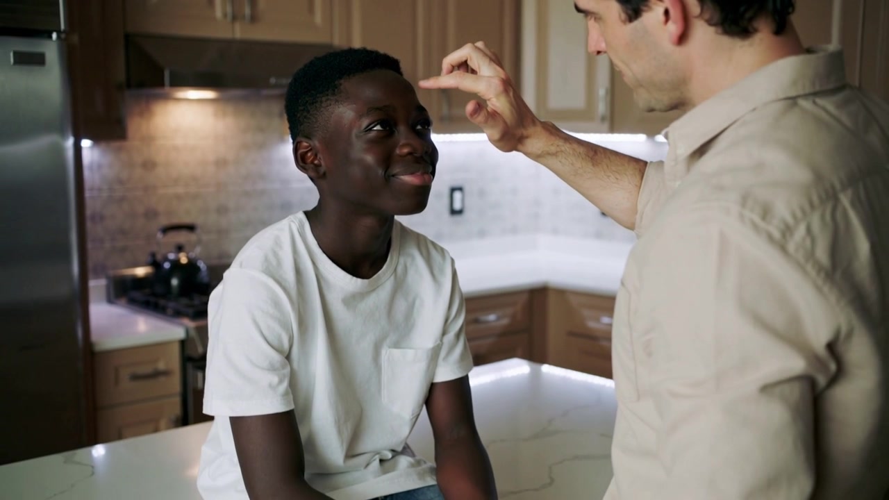 Leaning forward, adult in beige shirt touching boy in white tee at kitchen island, measuring fever