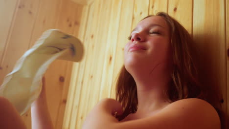 close up of woman seated next to sister fanning herself with felt hat inside warm steamy sauna room while engaging in soft conversation, surrounded by wooden walls and gentle mist atmosphere