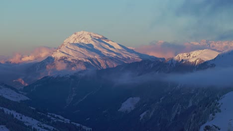 Fesselnde-Luftperspektive-Bietet-Einen-Faszinierenden-Blick-Auf-Den-Sonnenaufgang,-Der-Einen-Warmen-Glanz-Auf-Die-Silhouette-Einer-Schneebedeckten-Bergkette-Wirft,-Gefilmt-Von-Einer-Drohne