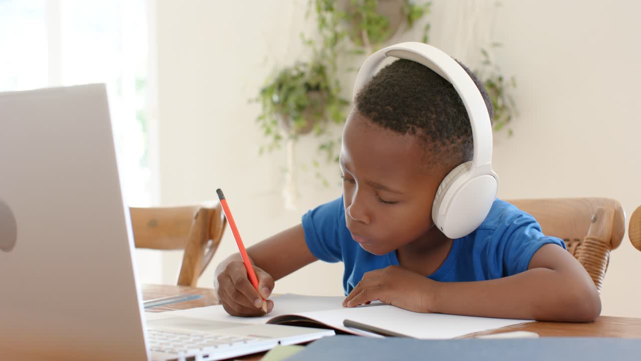 African American child writing notebook with pencil at home with headset raising hand to laptop cue