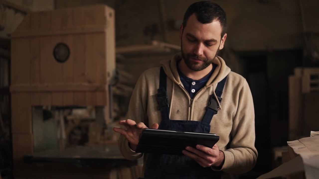 Young carpenter walking through the workshop with tablet in hands. Workshop with timber and equipment stuff on the background