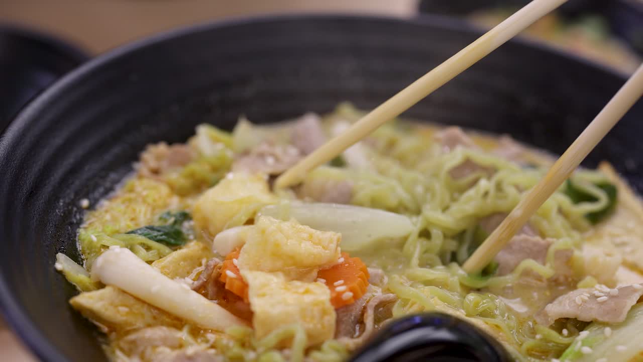 Hand uses chopsticks to lift green noodles from steaming Asian soup with vegetables and pork