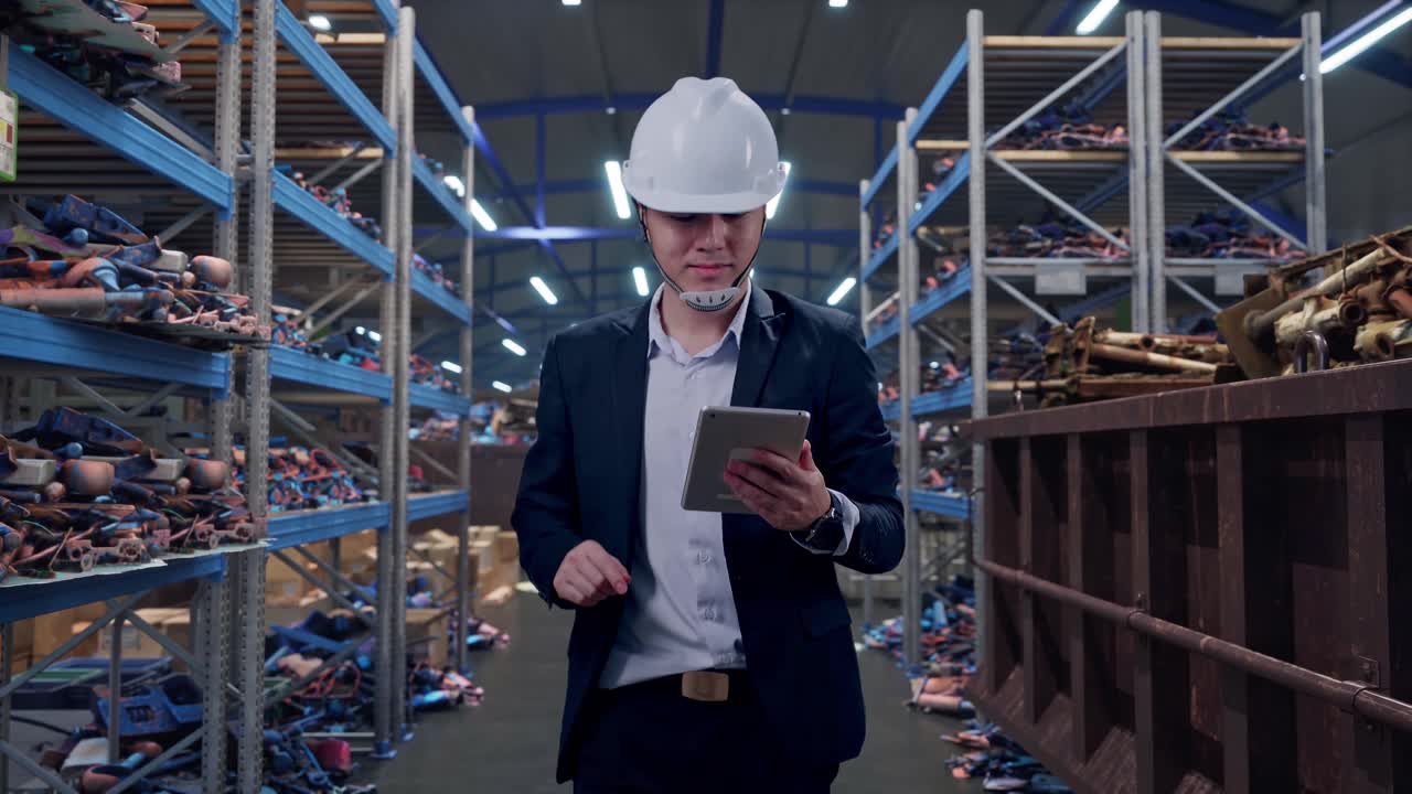 Uomo d'affari asiatico di fronte che usa un tablet mentre cammina in un magazzino industriale di rottami metallici e riciclaggio