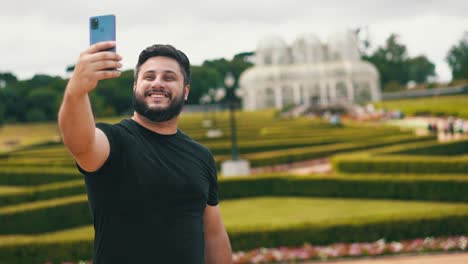 Junger-Mann-Lächelt-Und-Macht-Ein-Selfie-In-Einem-Wunderschönen-Botanischen-Garten-Im-Park-Mit-Einem-Großen-Gewächshaus-Im-Hintergrund