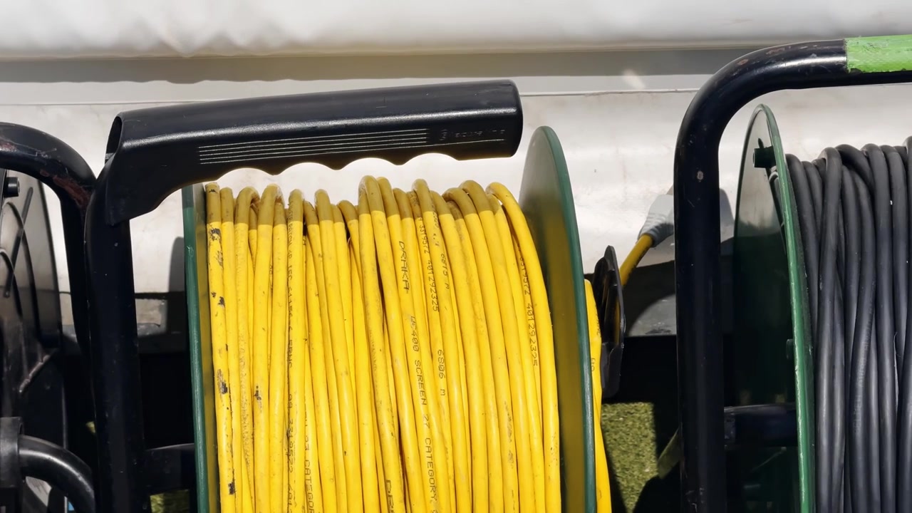 Close-up of yellow cable reel and coiled broadcast cables beside a sports court, showing event production equipment ready for setup.