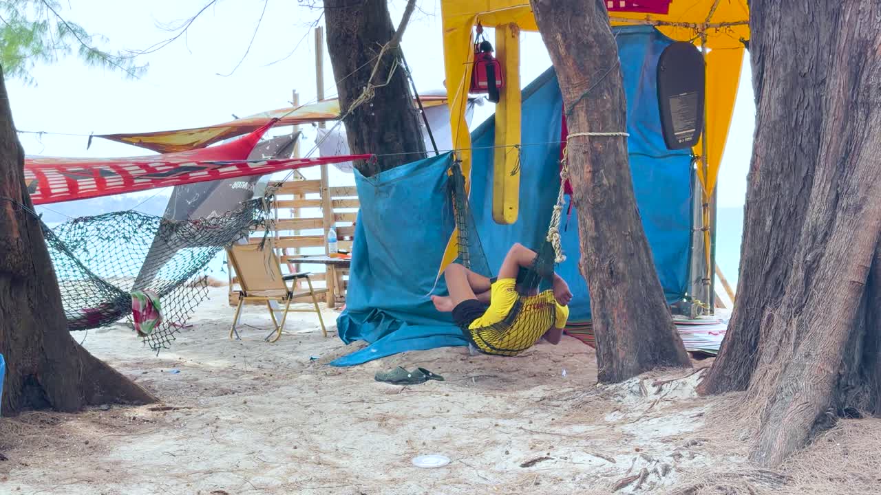En man vilar i en hängmatta under ett blått presenningsskydd på en sandstrand