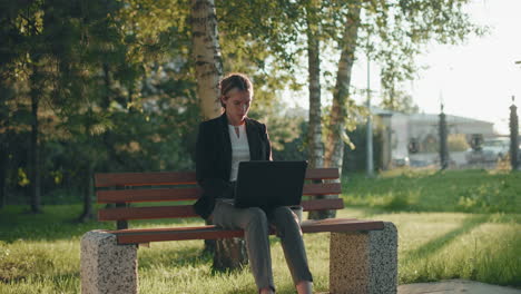 designer in blazer focused on design project using laptop seated in outdoor park during daylight with coffee cup placed beside, surrounded by greenery and natural light