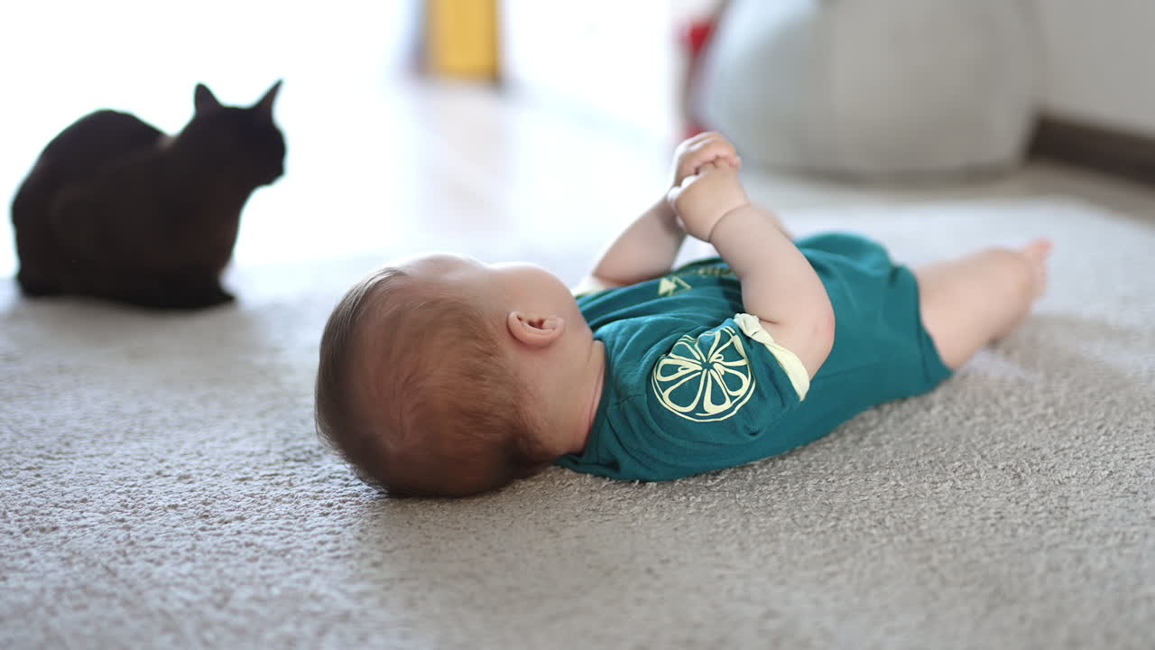 Calm peaceful baby lies on the floor. Black domestic cat sitting nearby the child. Blurred background.