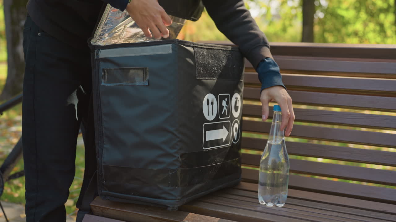 Un repartidor saca una bebida embotellada de una bolsa de reparto isotérmica colocada en un banco al aire libre y la deja sobre el banco. Manos enguantadas. Parque soleado. Entrega sin contacto. Listones de madera. Bolsa térmica negra de marca.