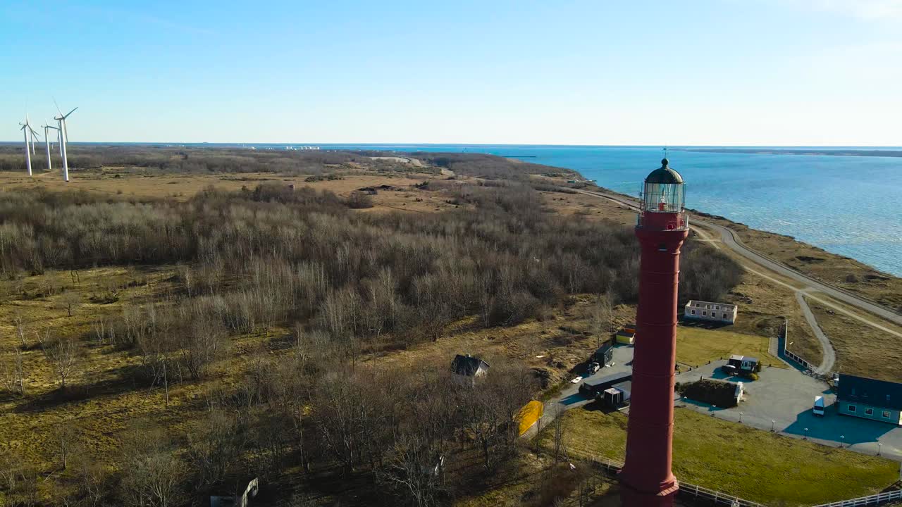 Splendide riprese aeree che volano attorno all'iconico faro rosso di Pakri sulla costa, rivelando un moderno parco eolico che genera energia rinnovabile in una giornata di sole a Paldiski, Estonia