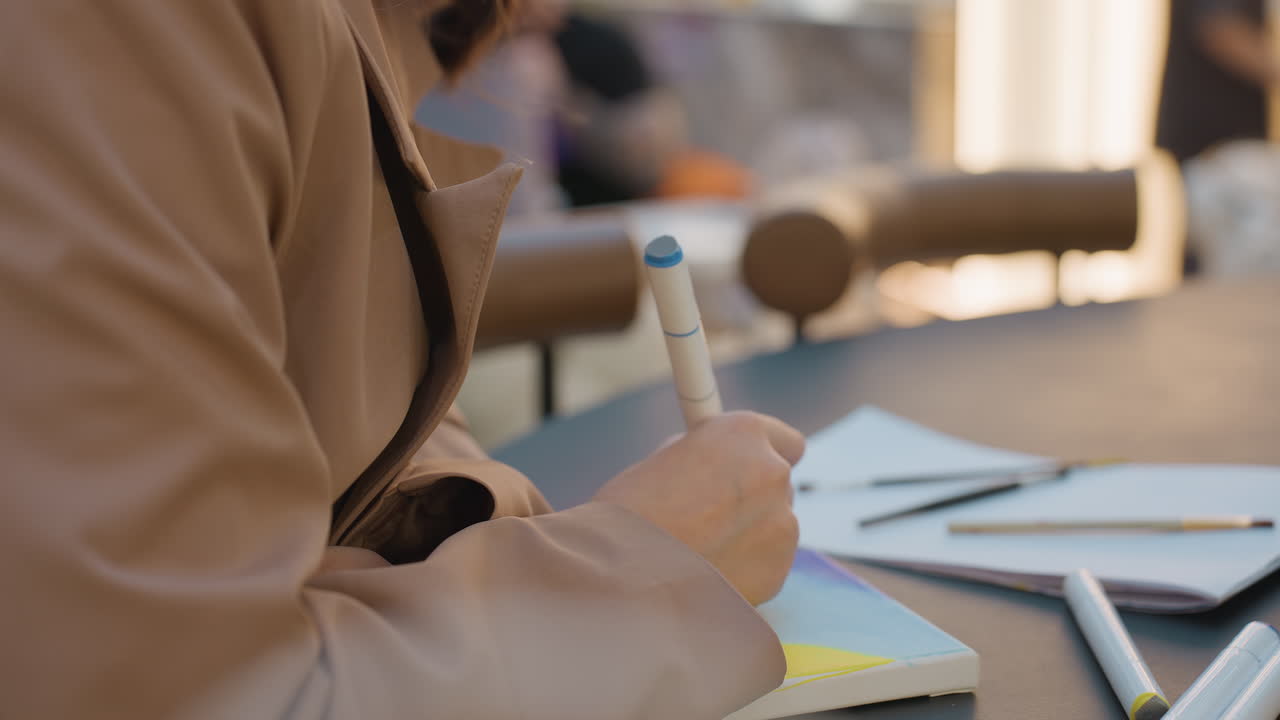 hands jotting notes, person taking notes at shared workspace, individual with pen and tablet at collaborative table, closeup of person composing notes at modern shared working environment