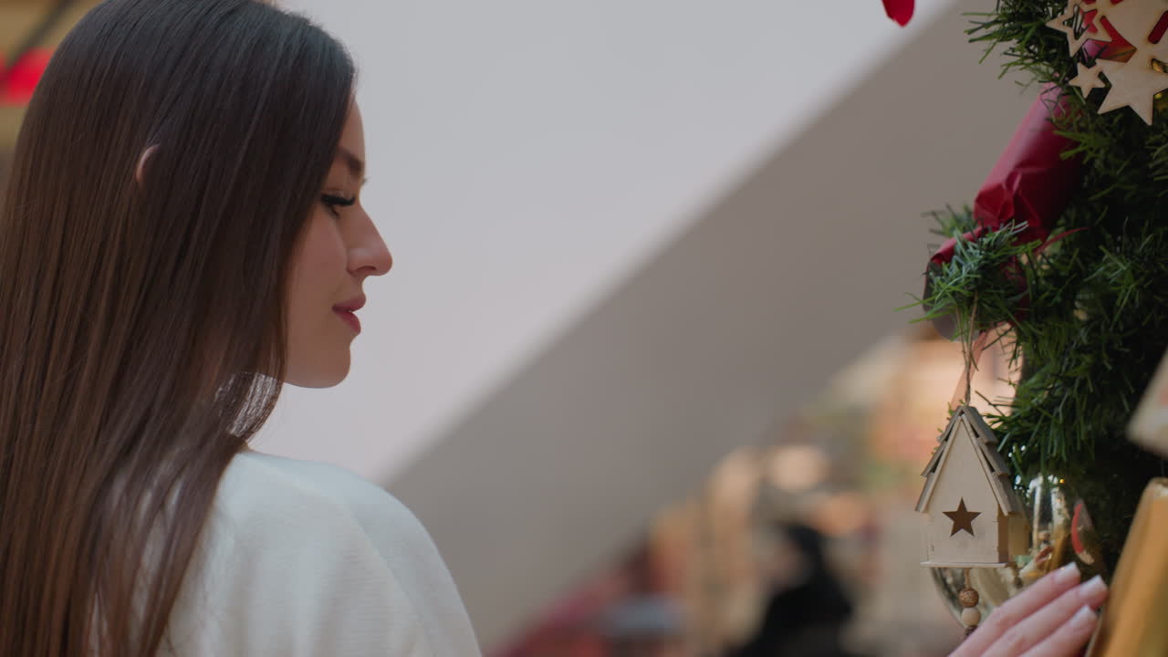 Woman walking around big Christmas tree, gently touching decorations with warm smile, enjoying holiday spirit in bustling mall with vibrant lights and background filled with shoppers