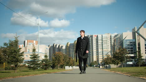 young man walking confidently on paved tarred road carrying bag, surrounded by vibrant trees, modern residential buildings, parked cars, and blurred figure in distant background under bright blue sky