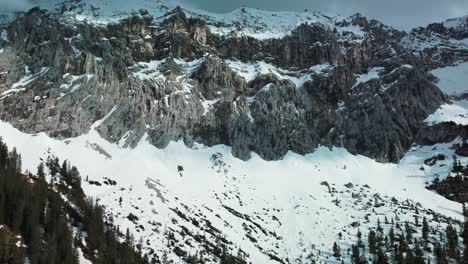 Wide-panorama-flight-at-a-glacier-rock-snow-mountain-top-near-Bavaria-Elmau-castle-in-the-Bavarian-Austrian-alps-on-a-cloudy-and-sunny-day-along-trees-and-forest-in-nature-with-avalanches-going-down