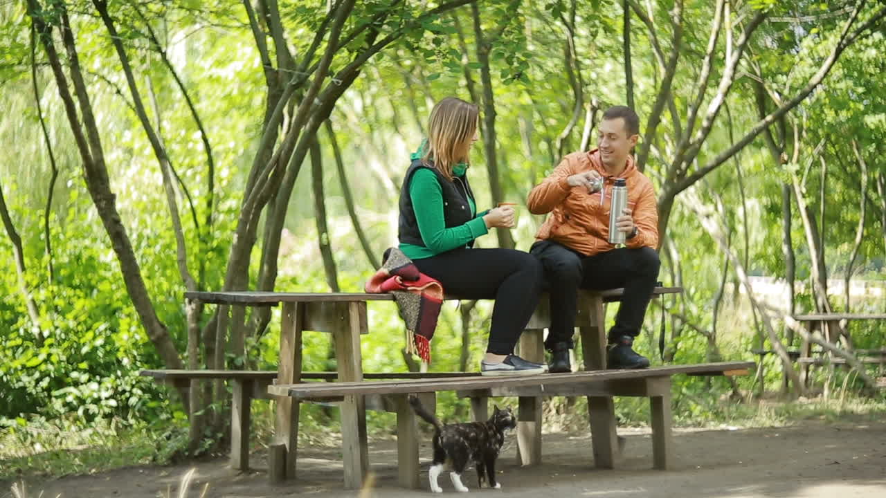 Couple Enjoying A Cup Of Tea. Happy young couple sitting on bench in park and drinking tea