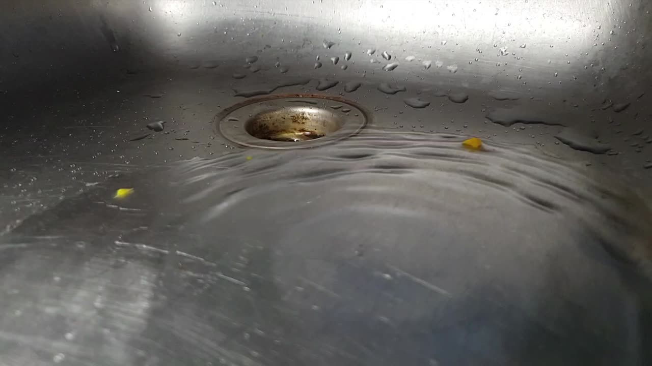 Faucet drainage hole in stainless kitchen sink with water dripping and splashing droplets in slow motion