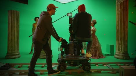 on film studio set shooting history movie green screen scene. moving cameraman on railway trolley shooting two costumed actors while director controls process. crew on big budget filmmaking