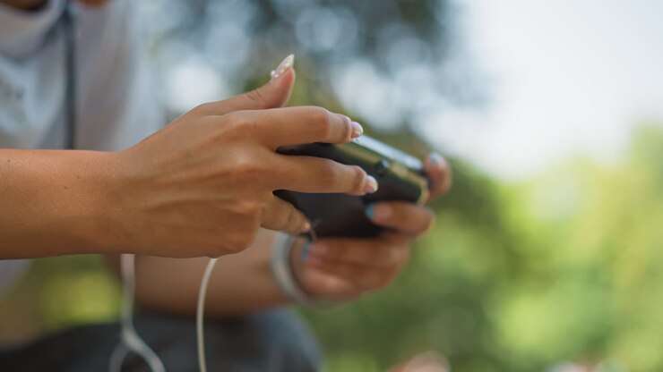 closeup hands holding mobile controller outdoors, focused player using earbuds and cable, sunlight filtering through trees, shallow depth of field, thumb pressing buttons with tense calm