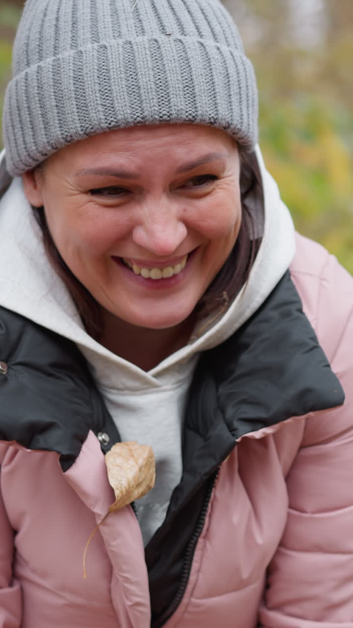 momento alegre como mujer en chaqueta rosa y gorra gris se ríe mientras las hojas de otoño caen a su alrededor, creando una escena de otoño lúdica y vibrante