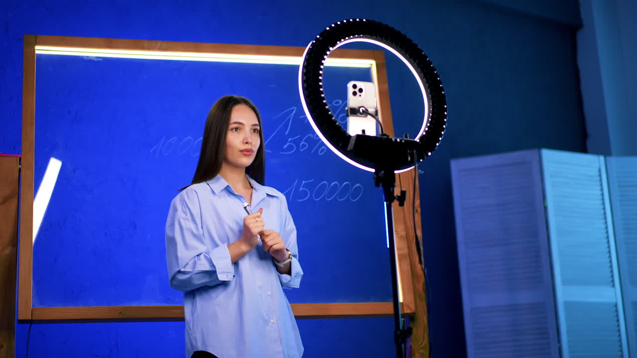 Young teacher in modern class. A young woman stands in front of a chalkboard, explaining a lesson while using a ring light to engage with her audience