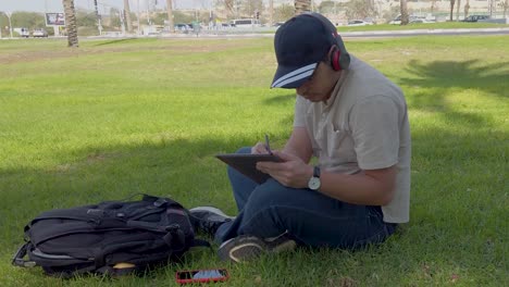 young man with headphones uses a tablet sitting on the grass in a park.
