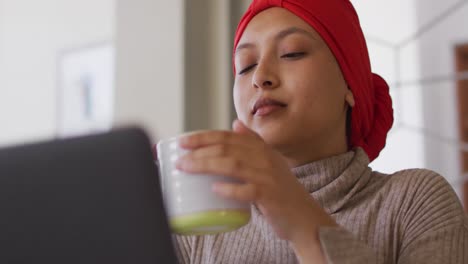 video of biracial woman in hijab sitting at desk at home working on laptop, drinking coffee