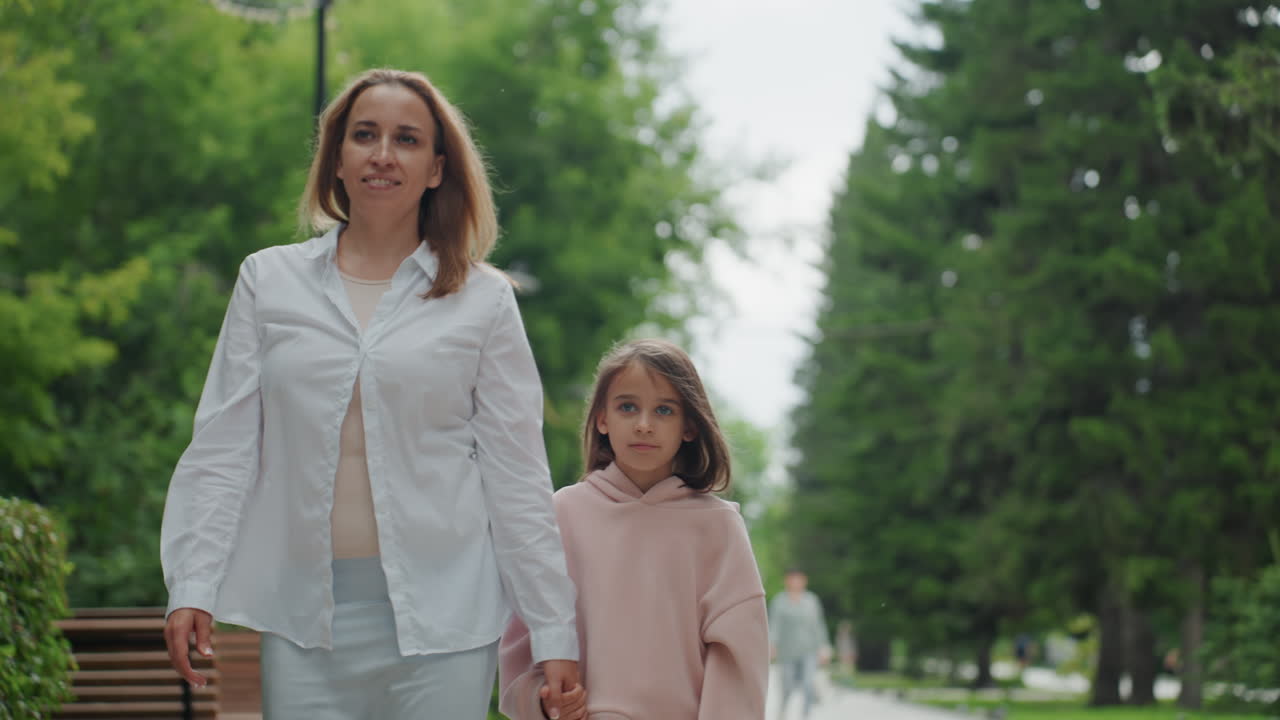 Mamma e figlia che camminano sotto alberi frondosi, conversazione calorosa e sorrisi, madre che guida la figlia lungo un sentiero lastricato, panchina e lampioni sullo sfondo, bagliore estivo, risate giocose, momento familiare urbano.