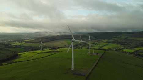 Wind-turbines-amidst-vibrant-green-fields,-with-rolling-hills-and-a-dramatic-cloudy-sky-in-the-backdrop,-aerial-view