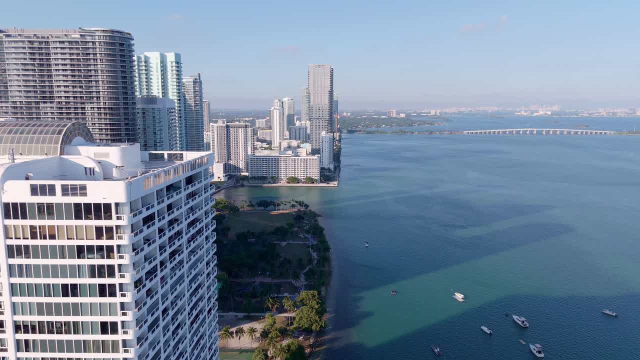 Aerial push-in view of Miami’s waterfront high-rises, a green city park, and the calm turquoise waters of Biscayne Bay with a scenic bridge in the background.