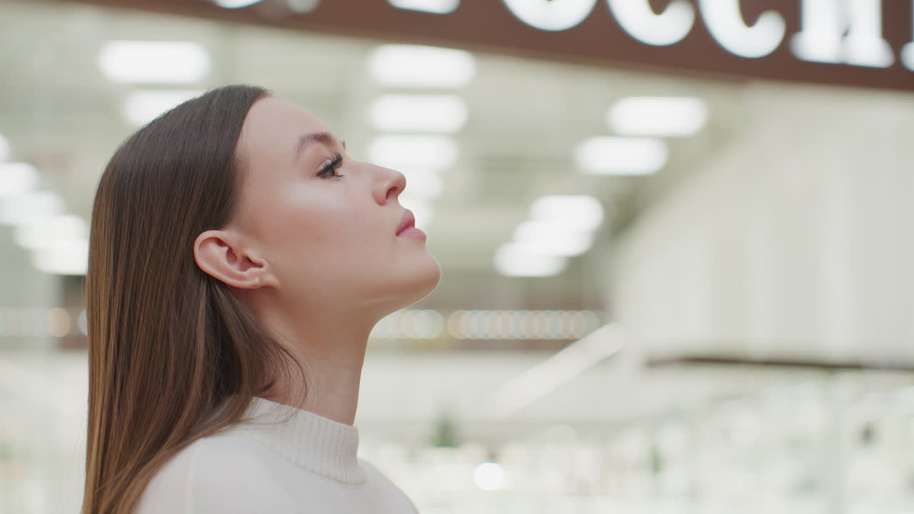 Calm lady walking up to Christmas tree, gazing at it peacefully while another shopper passes by in bright, well-lit mall, festive atmosphere with warm lighting, holiday decorations in background