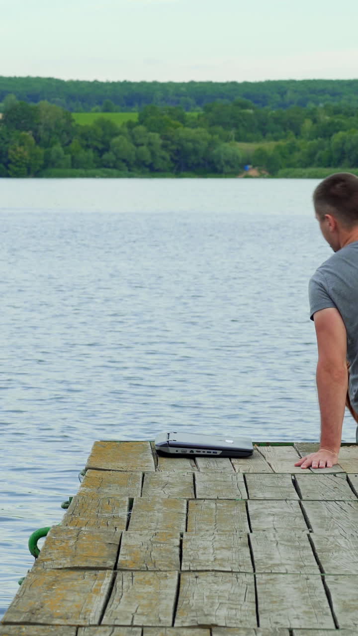 Man with his notebook is working outdoor at the lake. Old wooden pier. Vertical video