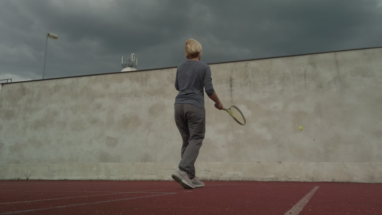 mujer golpea una pelota de tenis contra la pared bajo nubes de tormenta, ángulo bajo