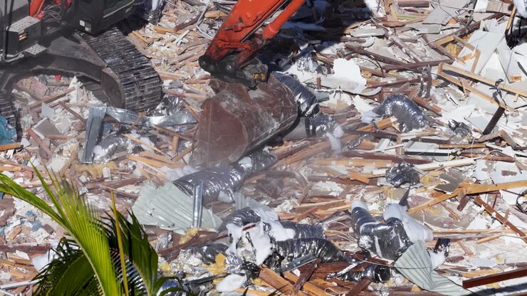 An excavator crushes building debris in a sunlit demolition site, with dust and rubble scattered. Close-up aerial view, steady camera, midday lighting