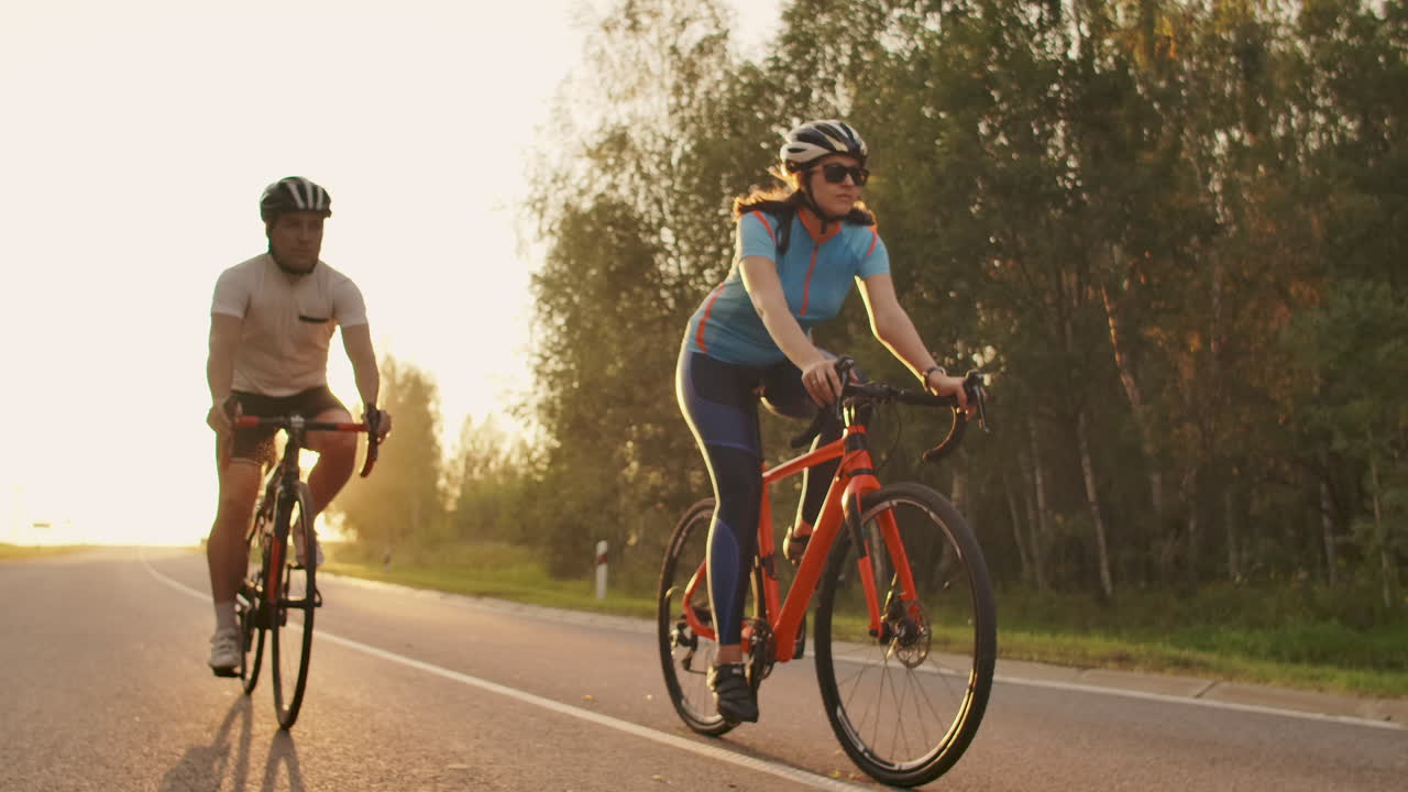 un hombre y una mujer montan bicicletas deportivas en la autopista al atardecer con engranajes y cascos protectores en cámara lenta 120 fps.