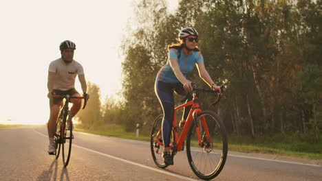 Un-Hombre-Y-Una-Mujer-Montan-Bicicletas-Deportivas-En-La-Carretera-Al-Atardecer-Con-Equipo-Y-Cascos-Protectores-En-Cámara-Lenta-A-120-Fps.