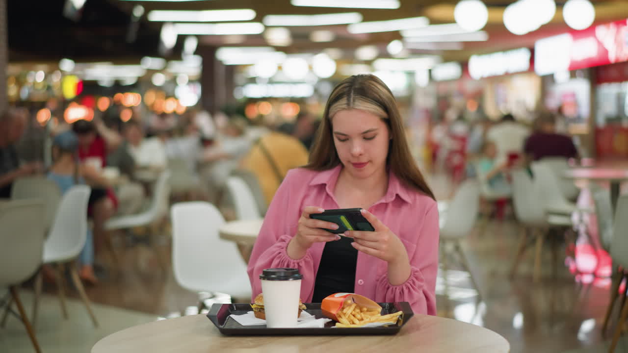 dama blanca en vestido rosa sentada en una mesa de madera en un restaurante ocupado, tomando una foto de su comida que consiste en una hamburguesa, papas fritas y una taza de café en una bandeja negra delante de ella