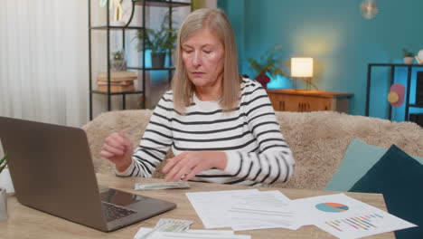 mature woman planning family budget on laptop reviewing documents counting money on table at home