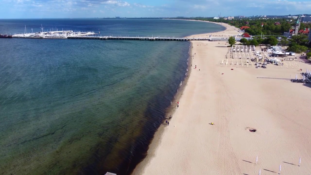 The coastline of Sopot, Poland, unfolds as a drone flies forward above the sandy beach, with the long pier and Baltic Sea in the background.